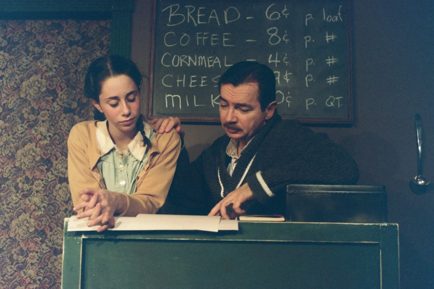 A woman and a man standing at a counter, reviewing a document. Behind them, a chalkboard displays prices for bread, coffee, cornmeal, cheese, and milk.