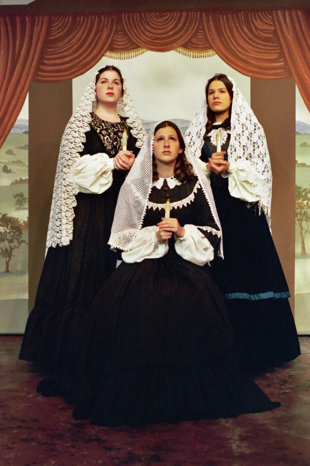Three women in historical costume with lace veils holding candles, standing in front of a backdrop with a painted scene. They appear to be part of a theatrical performance or period reenactment. The setting includes red curtains.