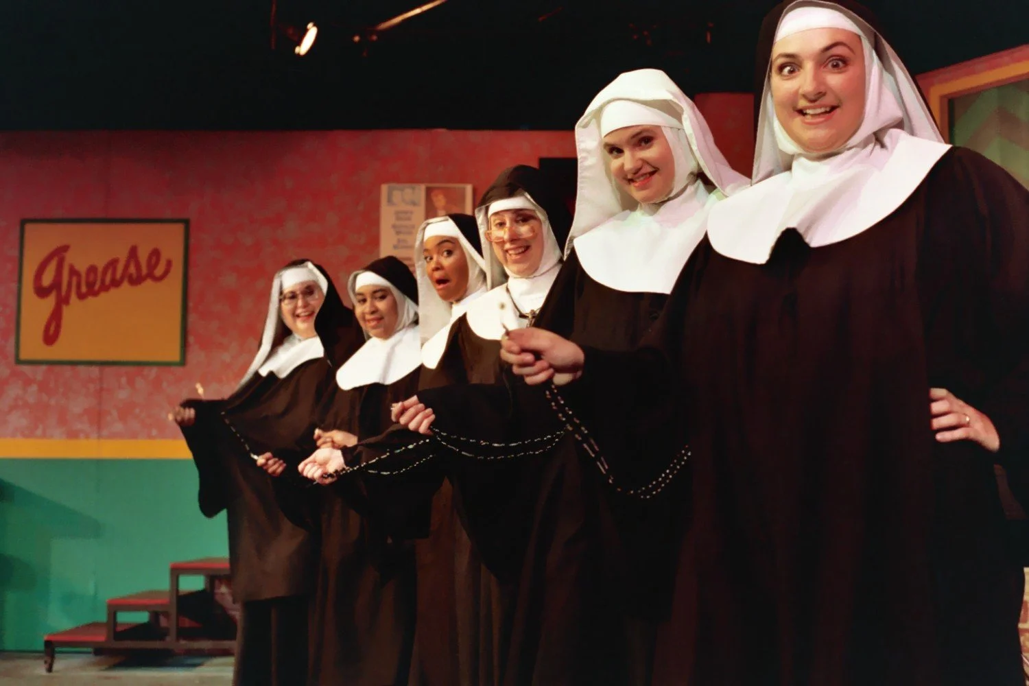 Group of women in nun costumes on stage holding rosary beads, with a "Grease" sign in the background.