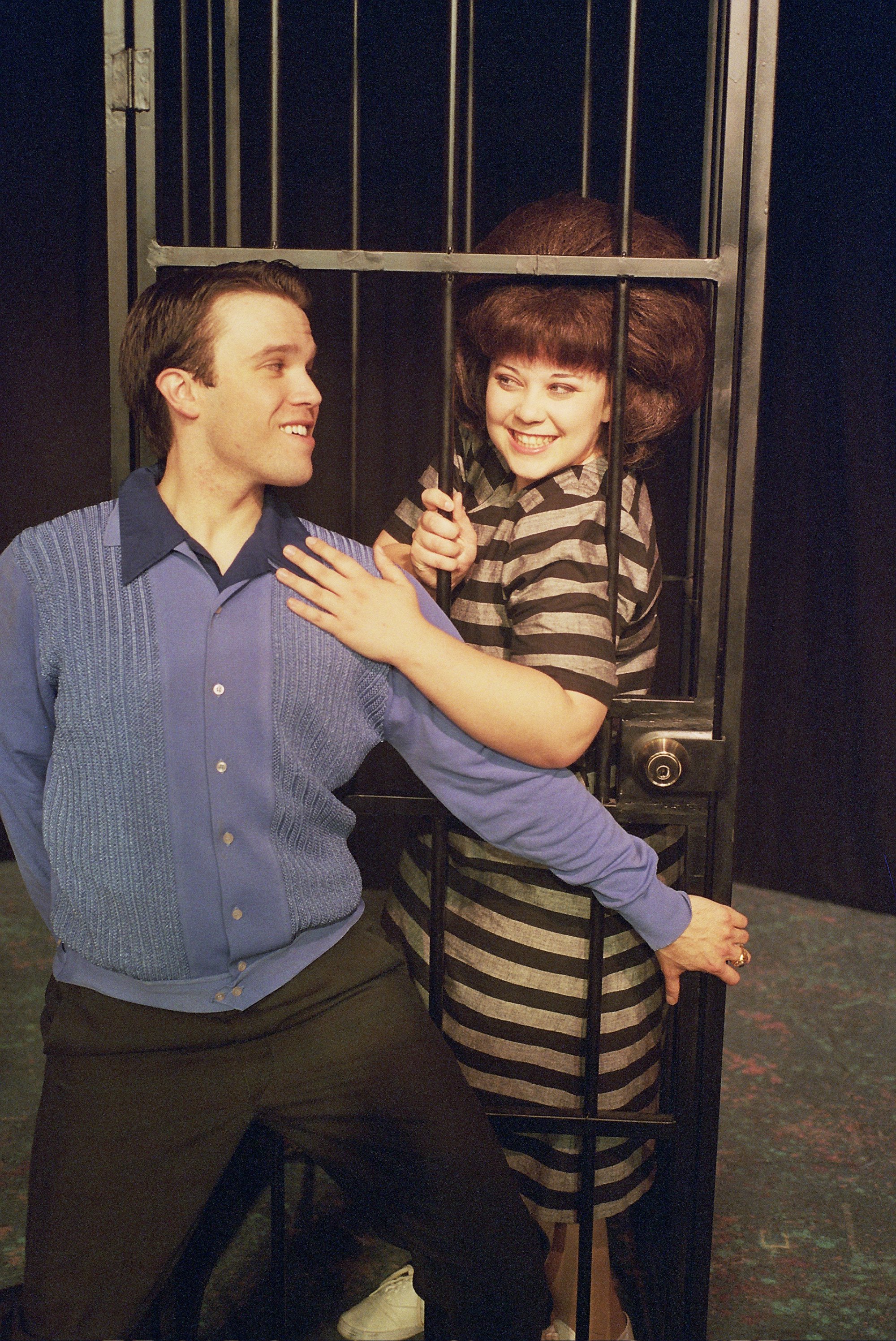 A man and a woman interacting playfully at a jail cell prop, with the woman inside the cell and the man outside. He is wearing a blue shirt and black pants, while she is in a striped dress. Both are smiling.