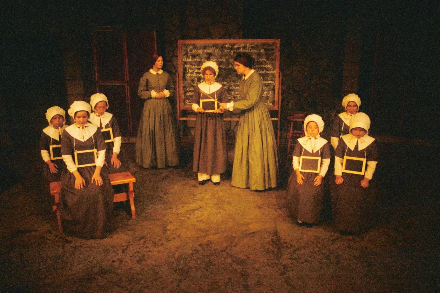 Group of people in historical attire, resembling a 19th-century schoolroom scene, with women and girls in bonnets and long dresses, seated around a chalkboard.