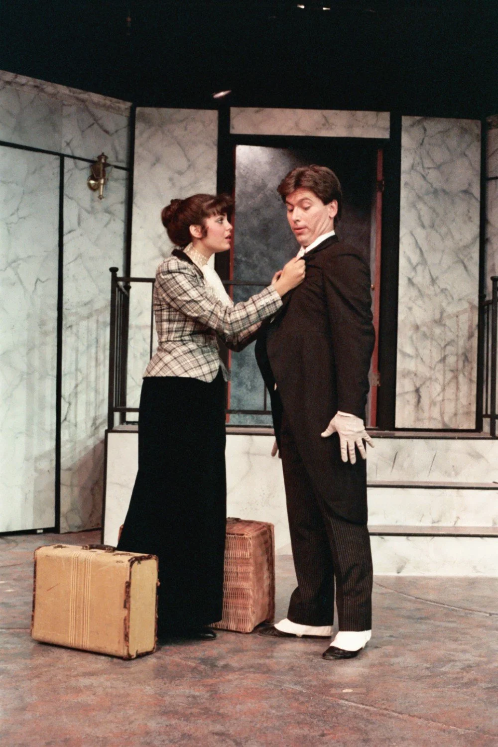Two actors on stage, one man wearing a suit and gloves, and a woman in a plaid jacket adjusting his tie, with suitcases on the floor