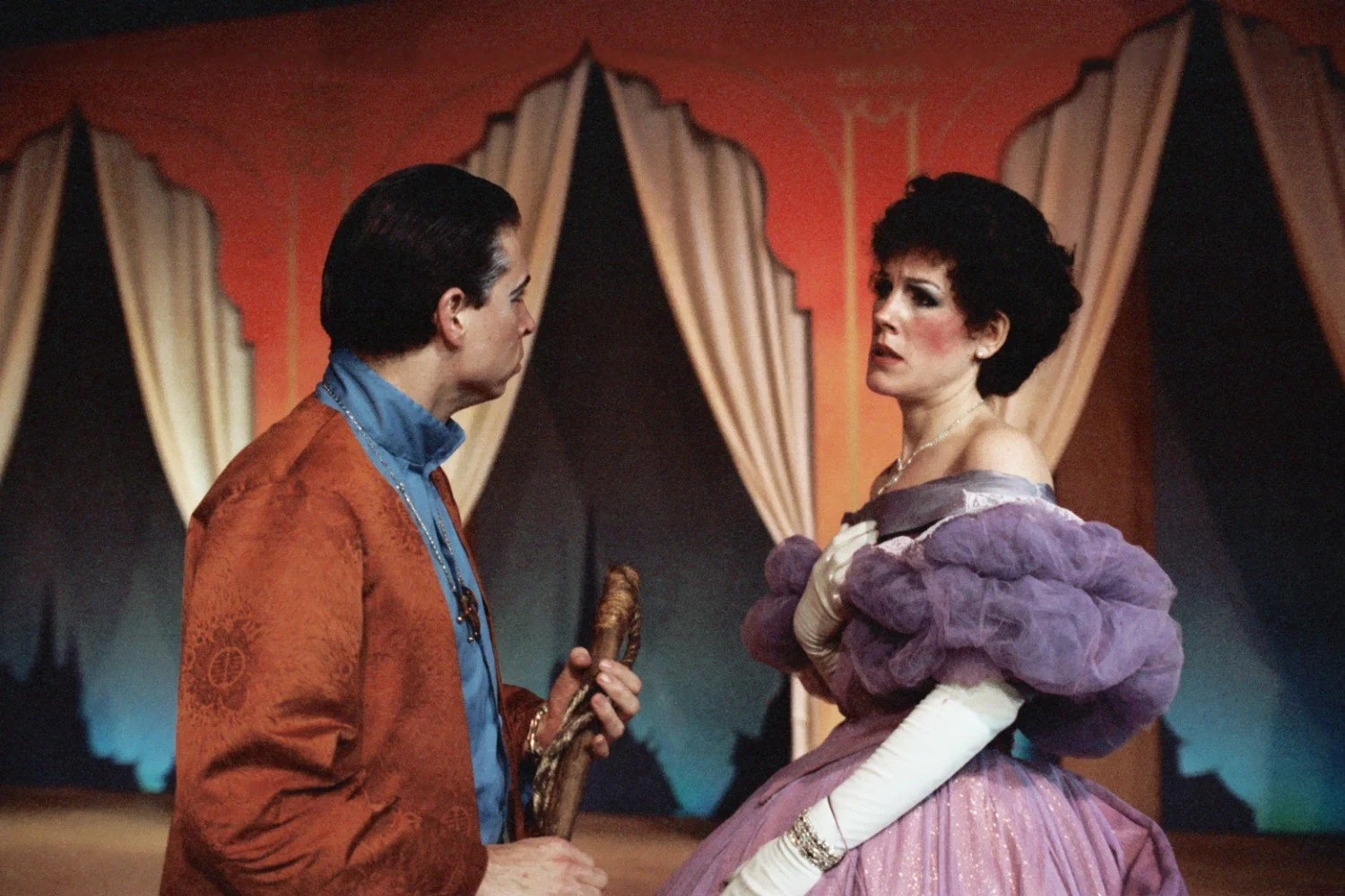 Two actors performing on stage, one male in a decorative red jacket and blue shirt holding a prop, and one female in a purple and white Victorian-style dress. The background features draped curtains and a painted set.