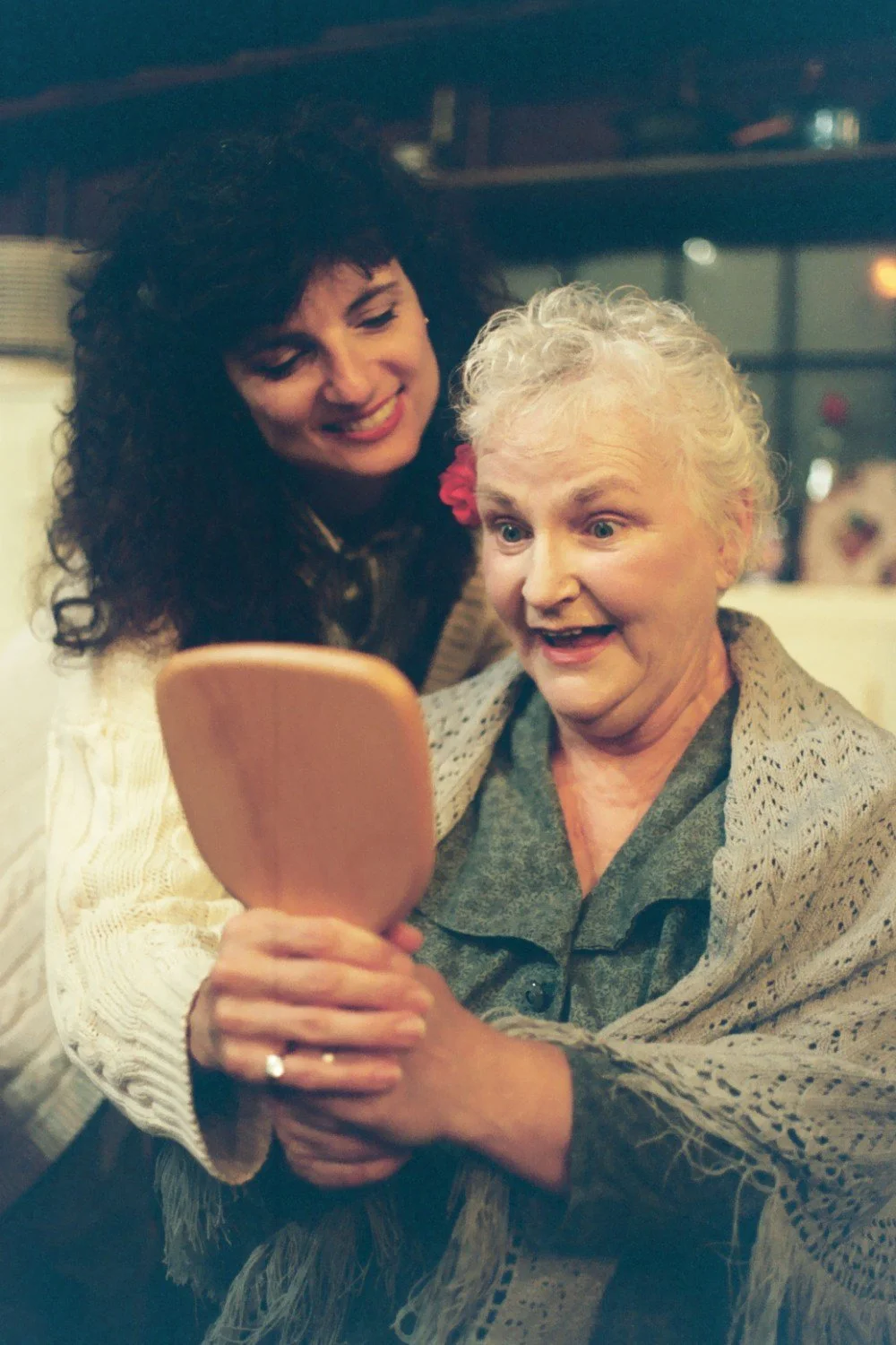 An elderly woman smiling while looking into a handheld mirror, assisted by a young woman standing behind her in a supportive gesture.