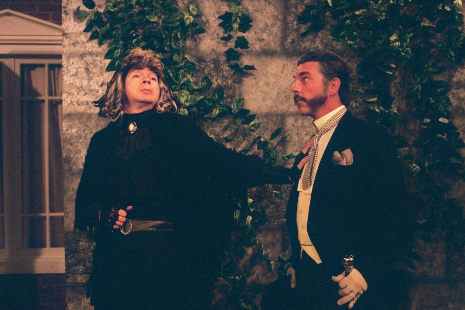 Two people dressed in costumes, possibly from a theatrical play. One is in a long dark robe with a brooch and a blond wig, the other in a formal suit with a pocket square and gloves. They are standing against a backdrop with climbing plants.
