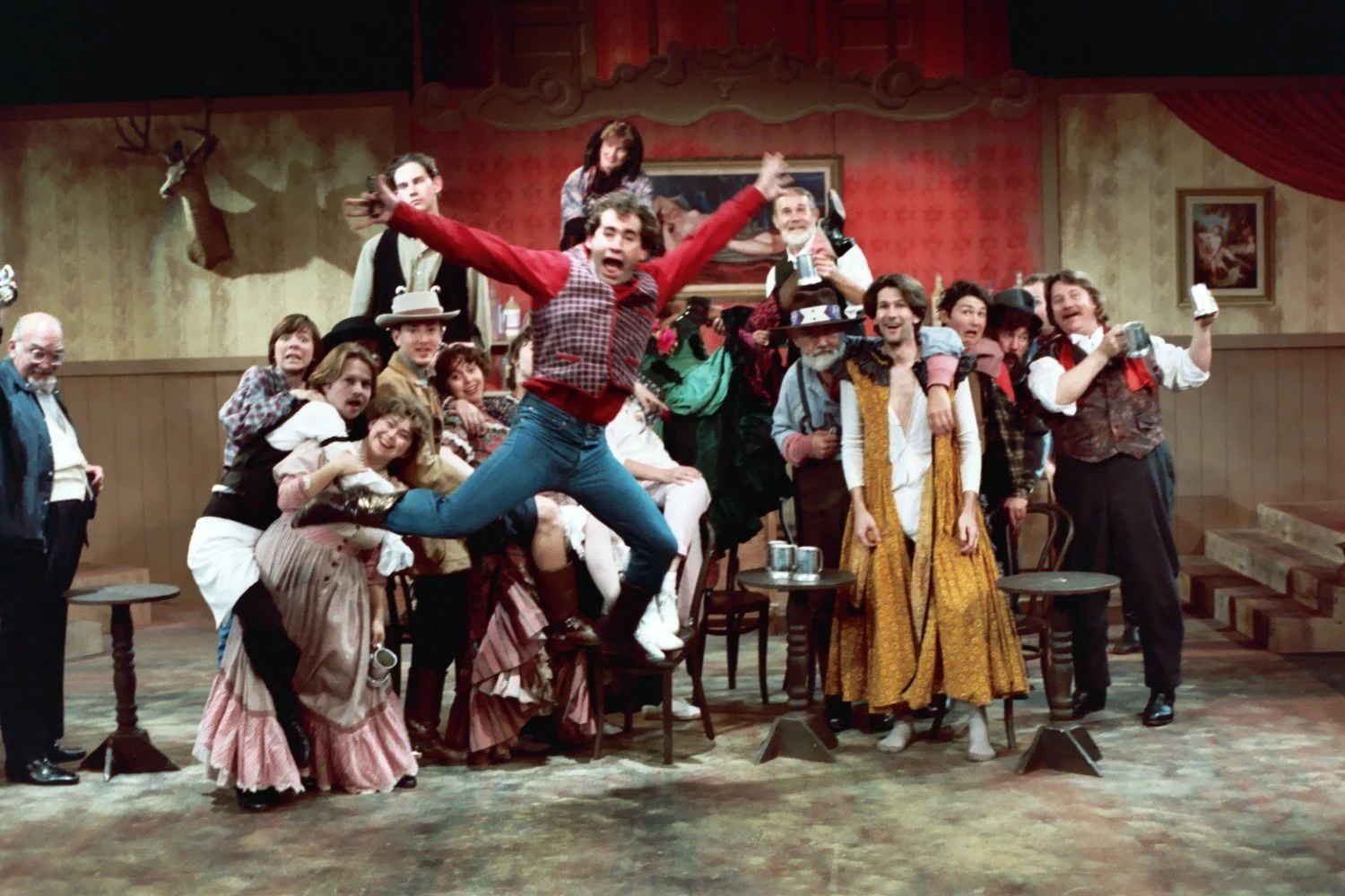 Performers in a theatrical production with a Western saloon theme, colorful costumes, and lively expressions, one jumping in the foreground.