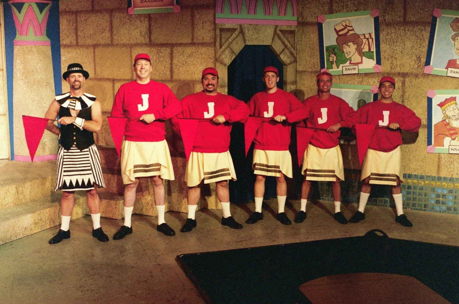 Group of six men in costumes on stage, five wearing red sweaters with the letter 'J' and matching skirts, holding red flags, one dressed in a striped outfit with a hat, colorful background with cartoon-style portraits.