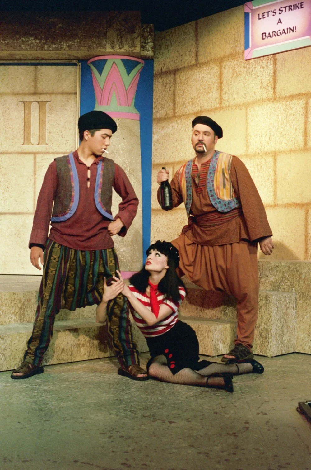 Three performers in theatrical costumes on stage; two men wearing vests, baggy pants, and berets, one holding a bottle; a woman in a striped shirt kneeling, holding one man's leg; backdrop with "Let's Strike a Bargain!" sign.