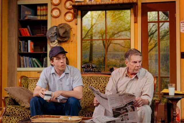 Two men sitting on a patterned sofa in a cozy room. The man on the left wears a cap and holds a cup, while the man on the right is reading a newspaper. Behind them are bookshelves and a window with a view of trees. A milk glass is on a small table be