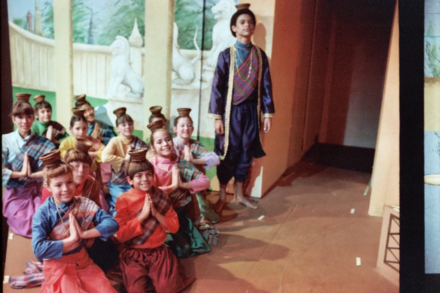 Children dressed in traditional Asian-style costumes performing a dance with hands held in prayer position and small baskets on their heads, standing on a stage with a painted backdrop of a palace setting.