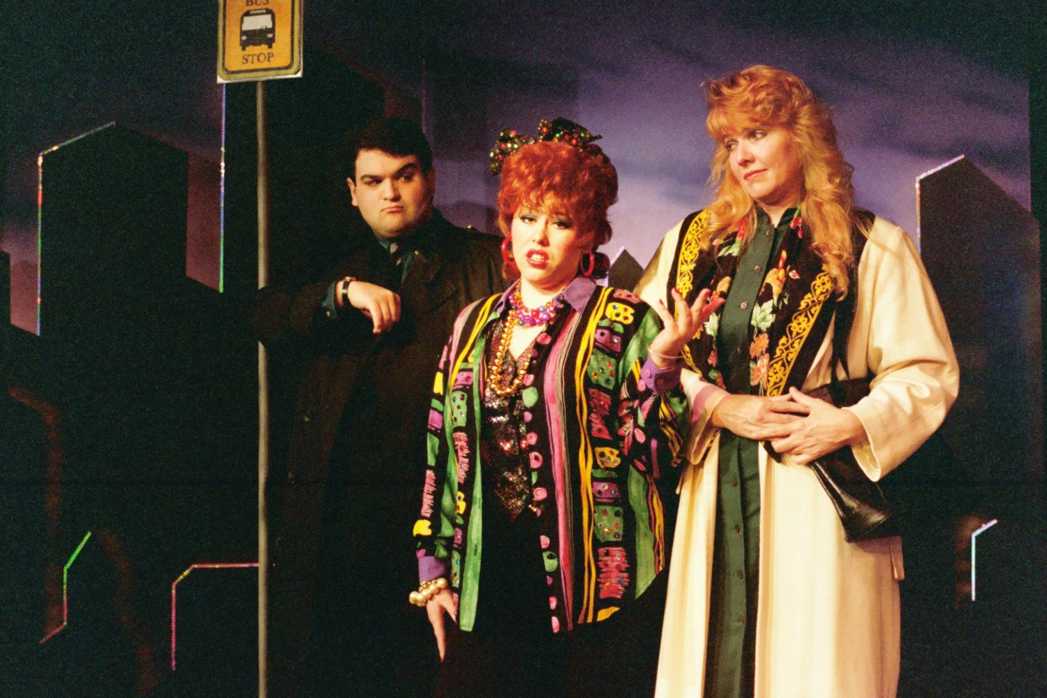 Three people standing at a bus stop at night, with a city skyline in the background. One person is wearing a colorful outfit, another a coat with an ornate scarf, and the third is in a dark suit.