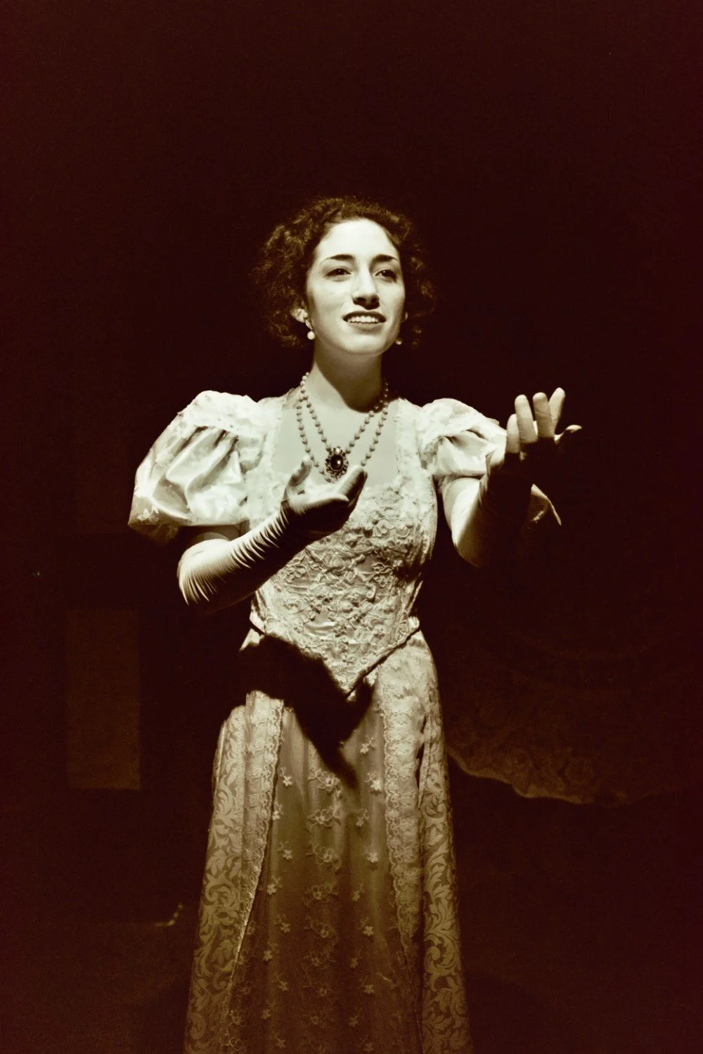 Sepia-toned image of a woman in vintage attire, wearing a lace dress, gloves, and a beaded necklace, gesturing expressively under spotlight.