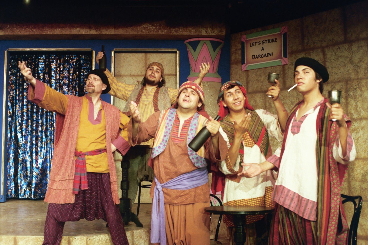 Group of actors in colorful period costumes on stage, holding bottles and cups, with a sign "Let's Strike a Bargain!"