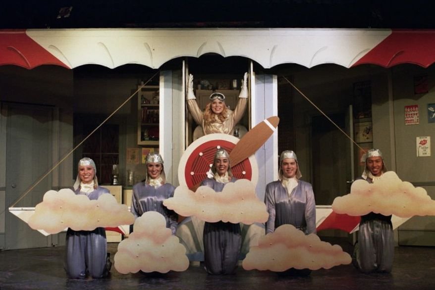 Stage performance with actors in vintage airplane costumes, wearing aviator hats and goggles, surrounded by cloud props.