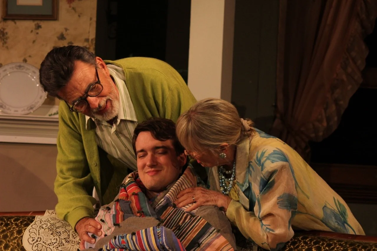 Two elderly individuals, one man and one woman, are leaning over a smiling young man wrapped in a colorful blanket. The older man is wearing glasses and a green sweater, while the older woman is in a floral blouse with pearls. They're indoors, probab