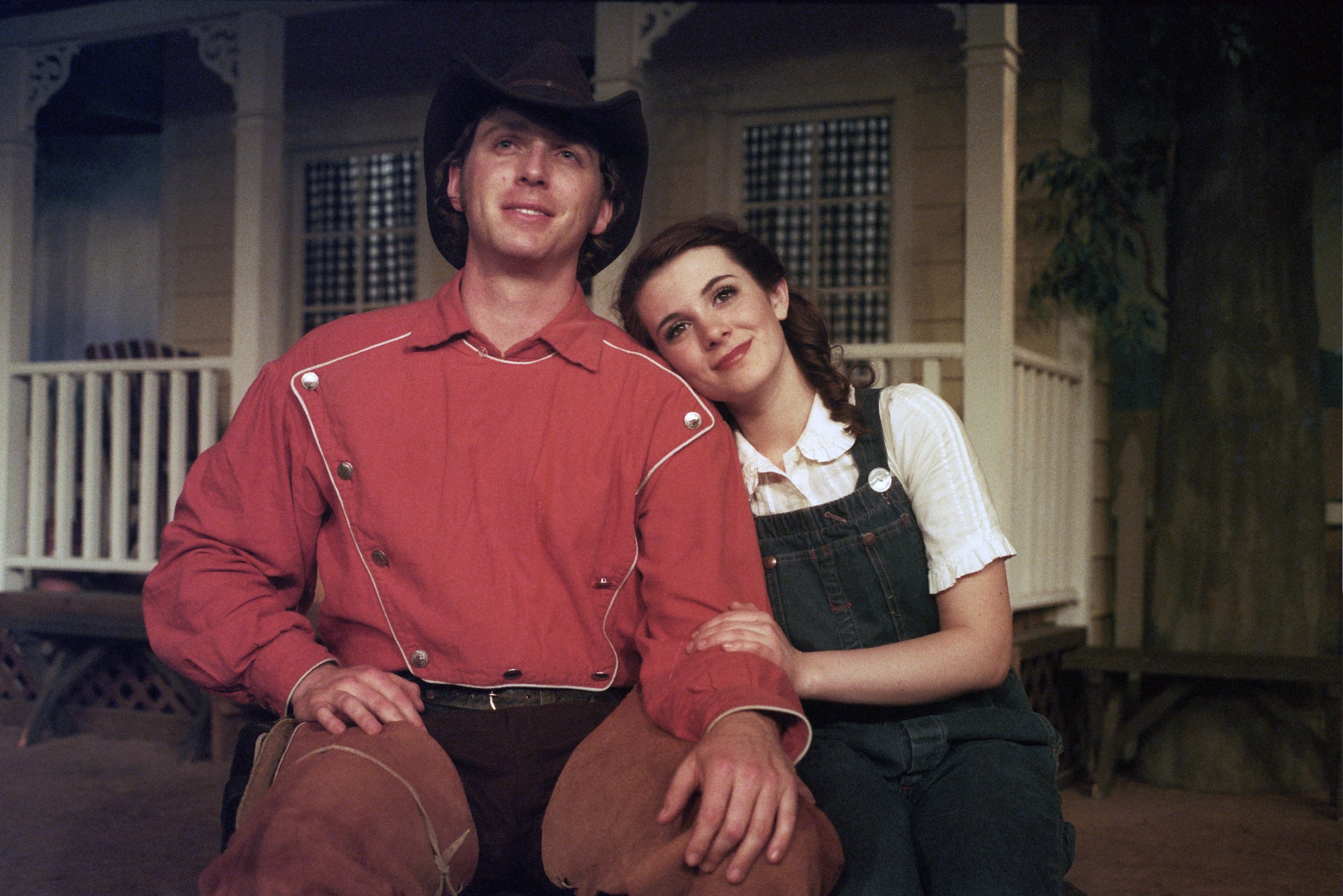 A man in a cowboy hat and red shirt sits next to a woman in overalls with her head resting on his shoulder. They appear to be in a theatrical stage setting with a house porch in the background.