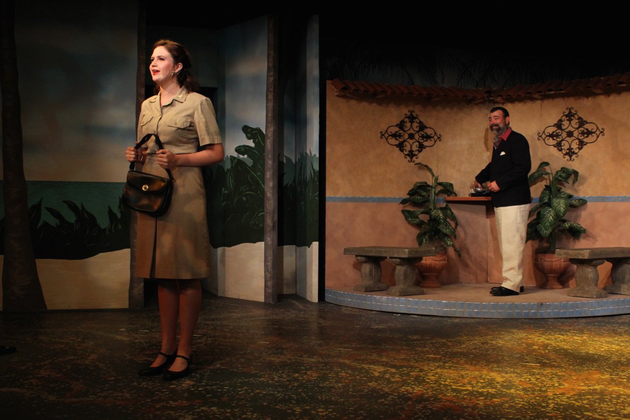Two actors performing on stage in a theatrical production with a tropical-themed set; a woman in a khaki dress holding a purse and a man in a black blazer and white pants. The backdrop includes a painted ocean scene and decorative wall elements.