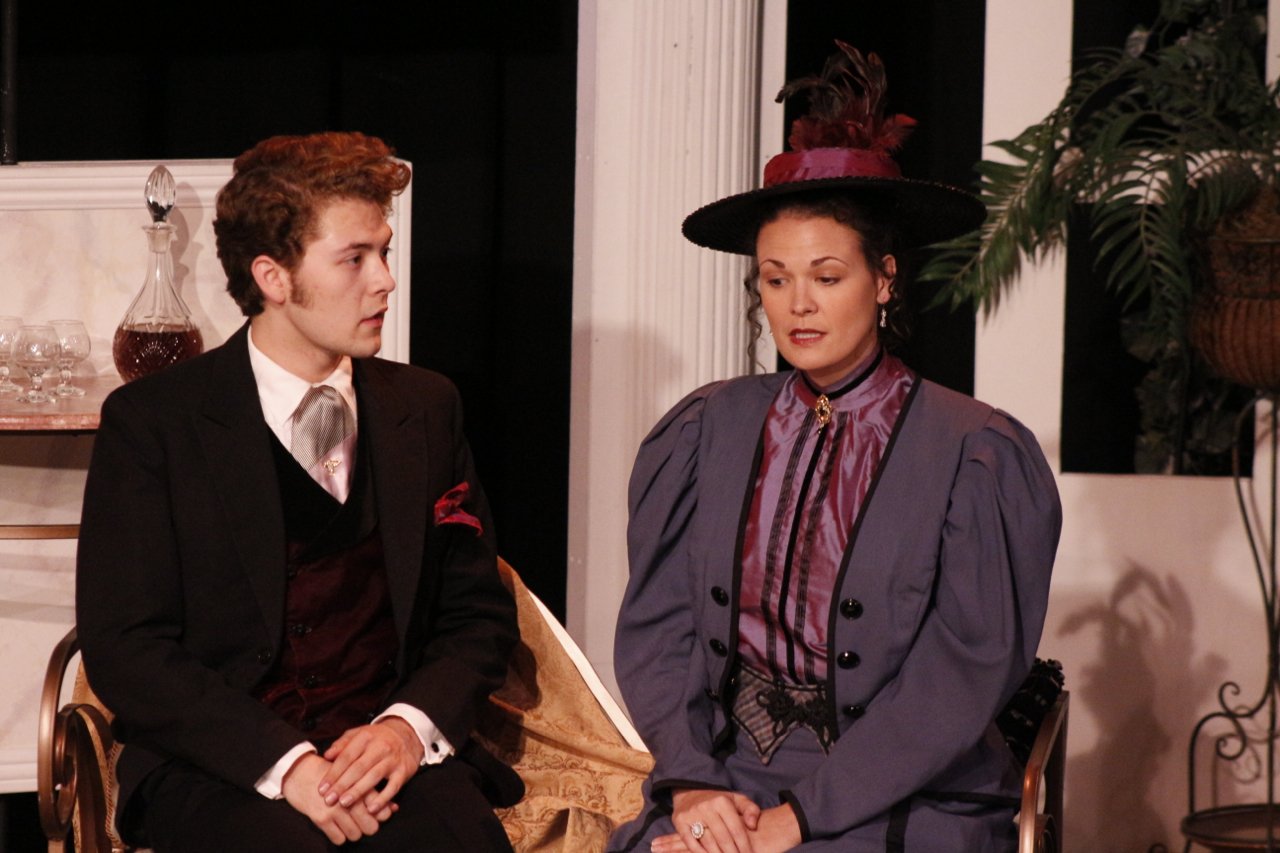 Two people in Victorian costumes sitting, one in a suit and the other in a purple dress and hat, with a decorative decanter and greenery in the background.