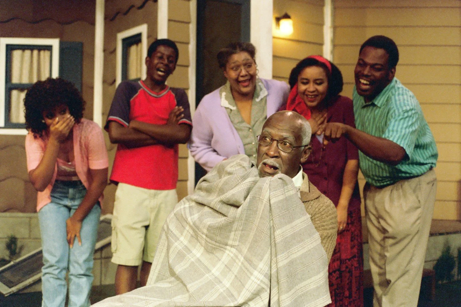 Group of people laughing and pointing at surprised man wrapped in blanket outside a house.