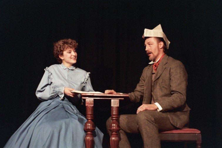 Two actors in period costumes perform on stage with a small table between them, one wearing a paper hat.