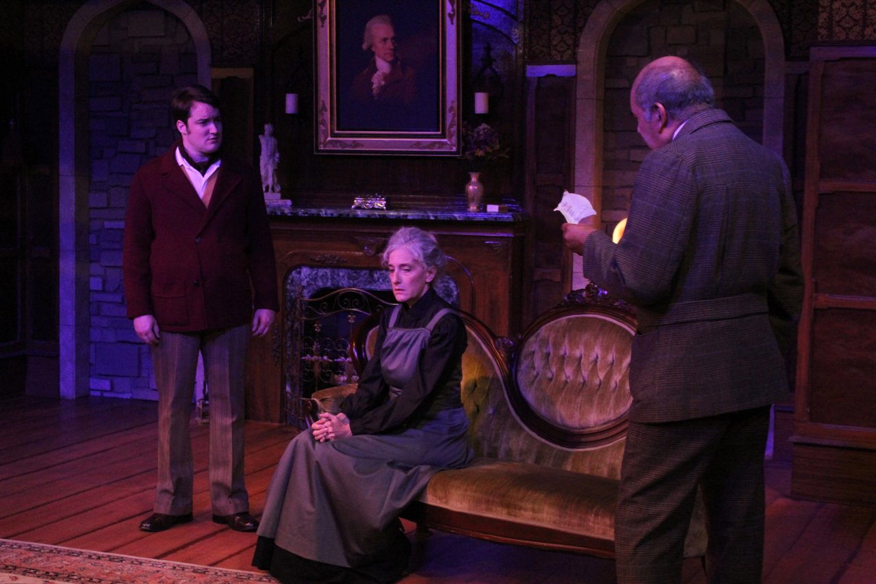Three actors perform a dramatic scene on stage in a Victorian-style setting with a fireplace and portrait in the background.