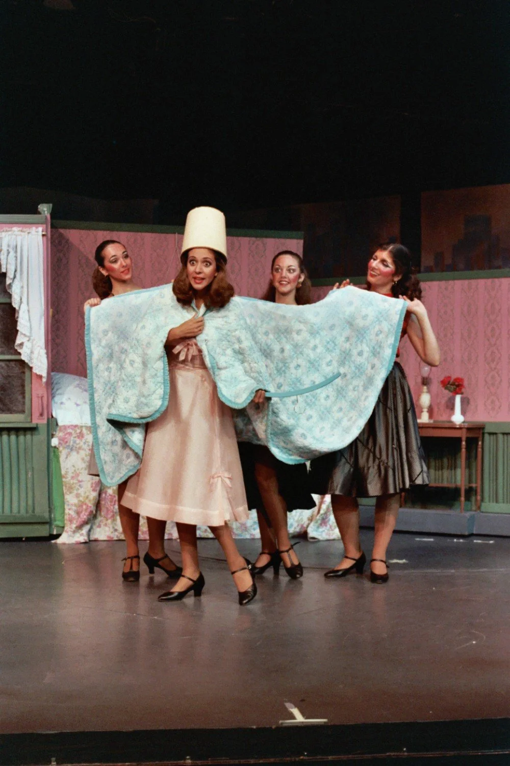 Four women performing on stage in a theatrical production, with one in a pink dress and hat, wrapped in a blue quilt, and the others holding it open.