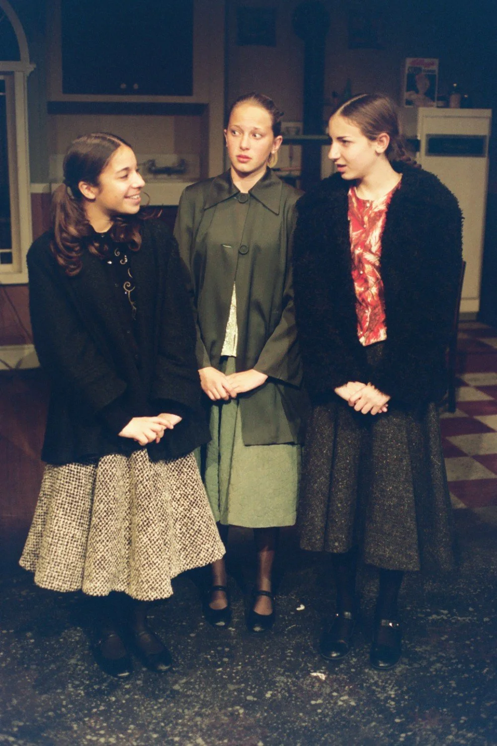 Three women standing in a kitchen setting, wearing vintage-style clothing. They are engaged in conversation, with one woman in the center looking surprised or confused.