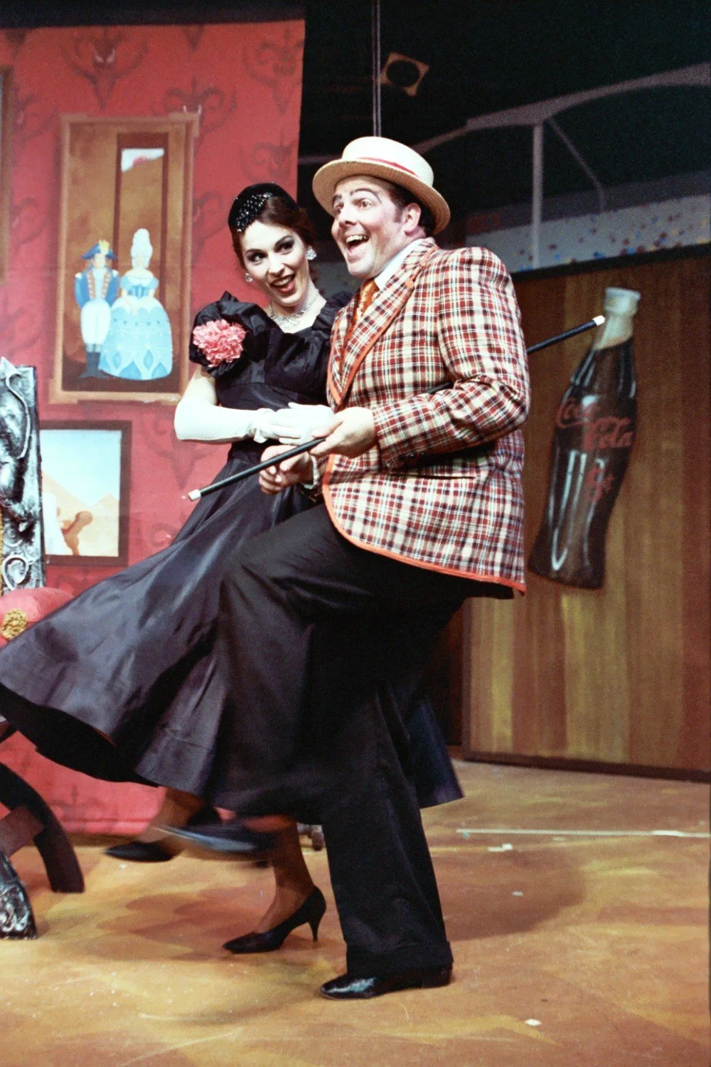 Two actors performing on stage in vintage attire with a painted backdrop showing historical costumes; a Coca-Cola bottle prop in the background.