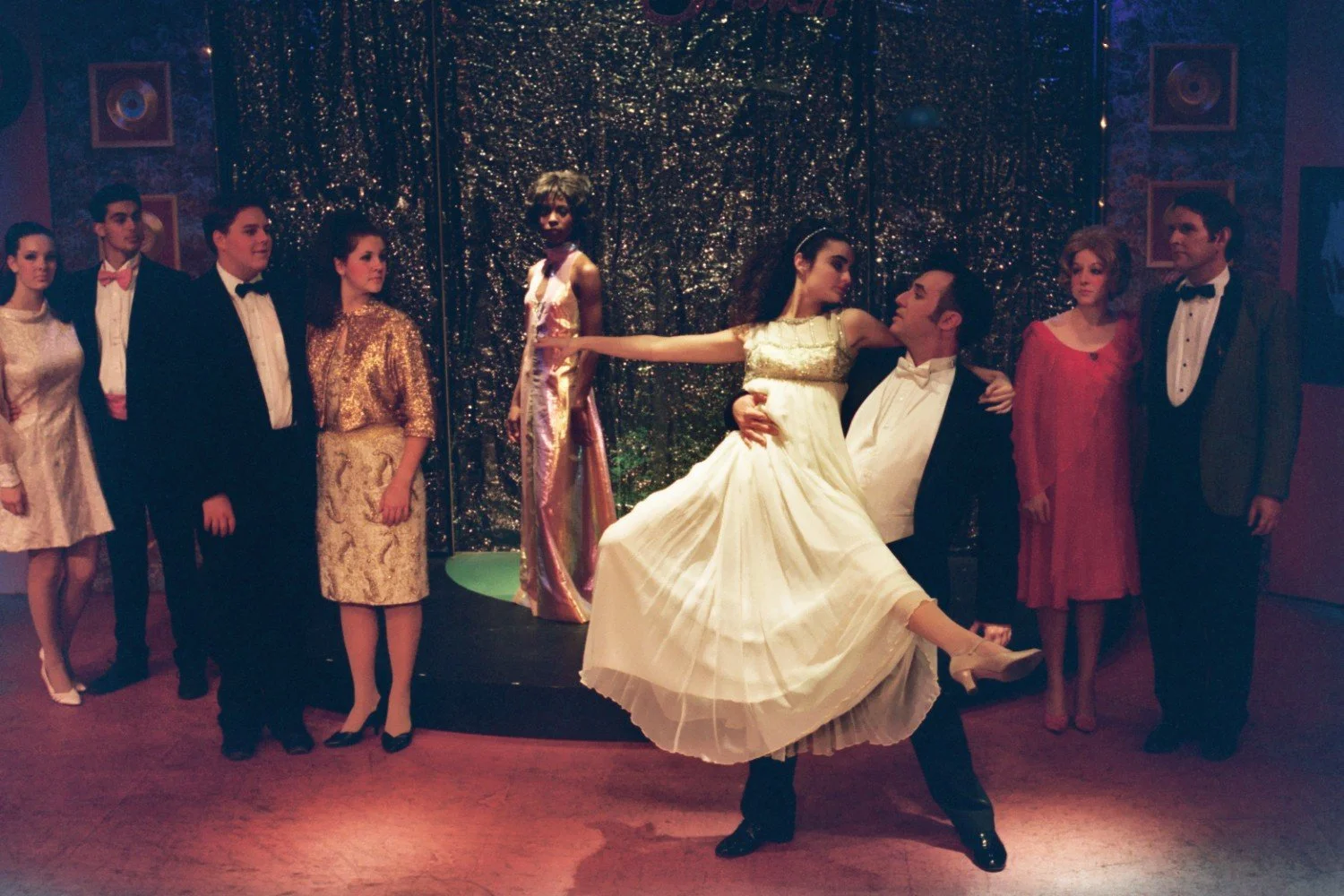 A group of people dressed in formal attire, gathered in front of a glittering backdrop. In the center, a man in a tuxedo holds a woman in a white dress as if they are dancing. Other individuals stand nearby, watching the couple.