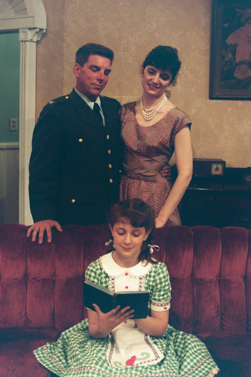 A man in a military uniform and a woman in a vintage dress stand behind a young girl sitting on a red sofa reading a book.