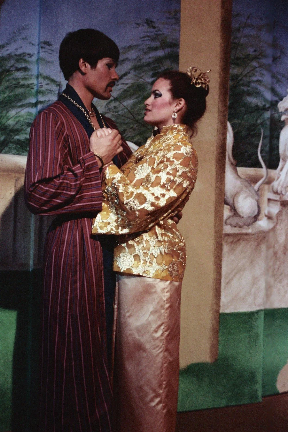 A man and a woman in theatrical costumes stand close together, holding hands, in front of a painted backdrop with foliage and architectural elements.