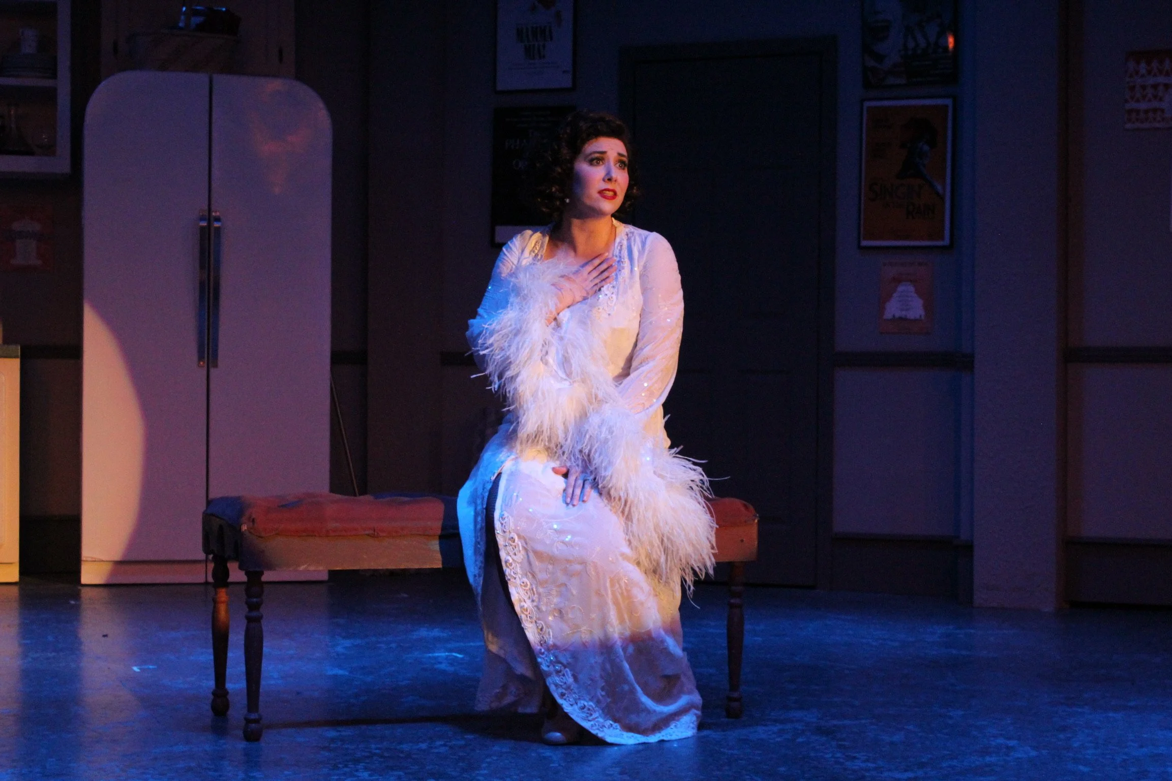 A woman in a white, feathered gown sits on a bench on stage, looking emotional. The background includes a refrigerator and framed posters. Stage lighting casts a dramatic effect.
