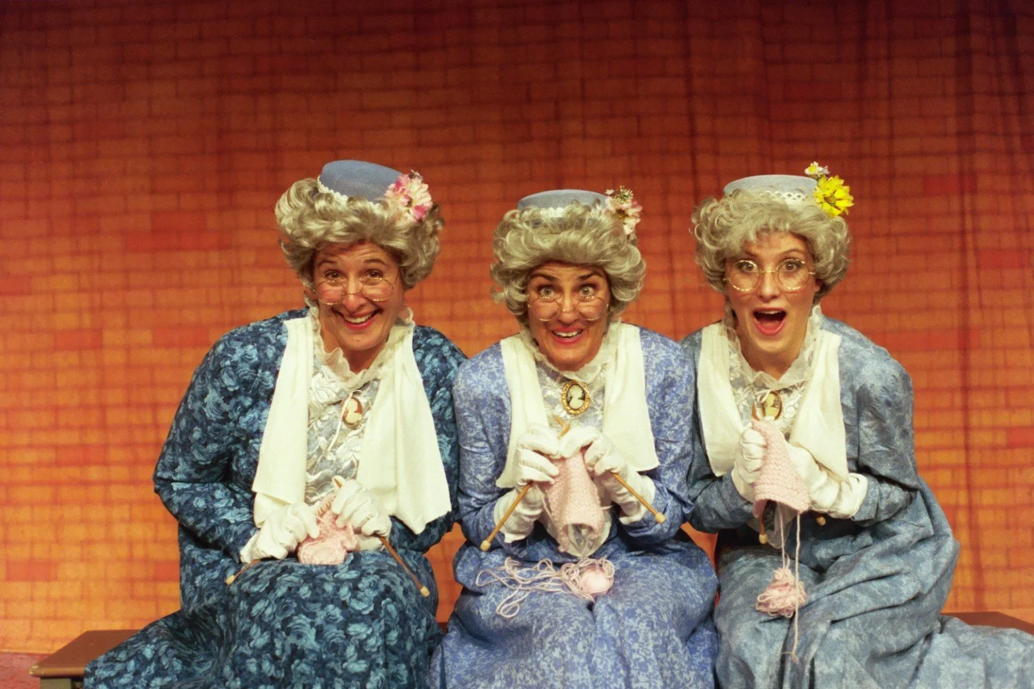 Three people dressed as elderly women in blue dresses and wigs, sitting and knitting on stage with an orange brick background.