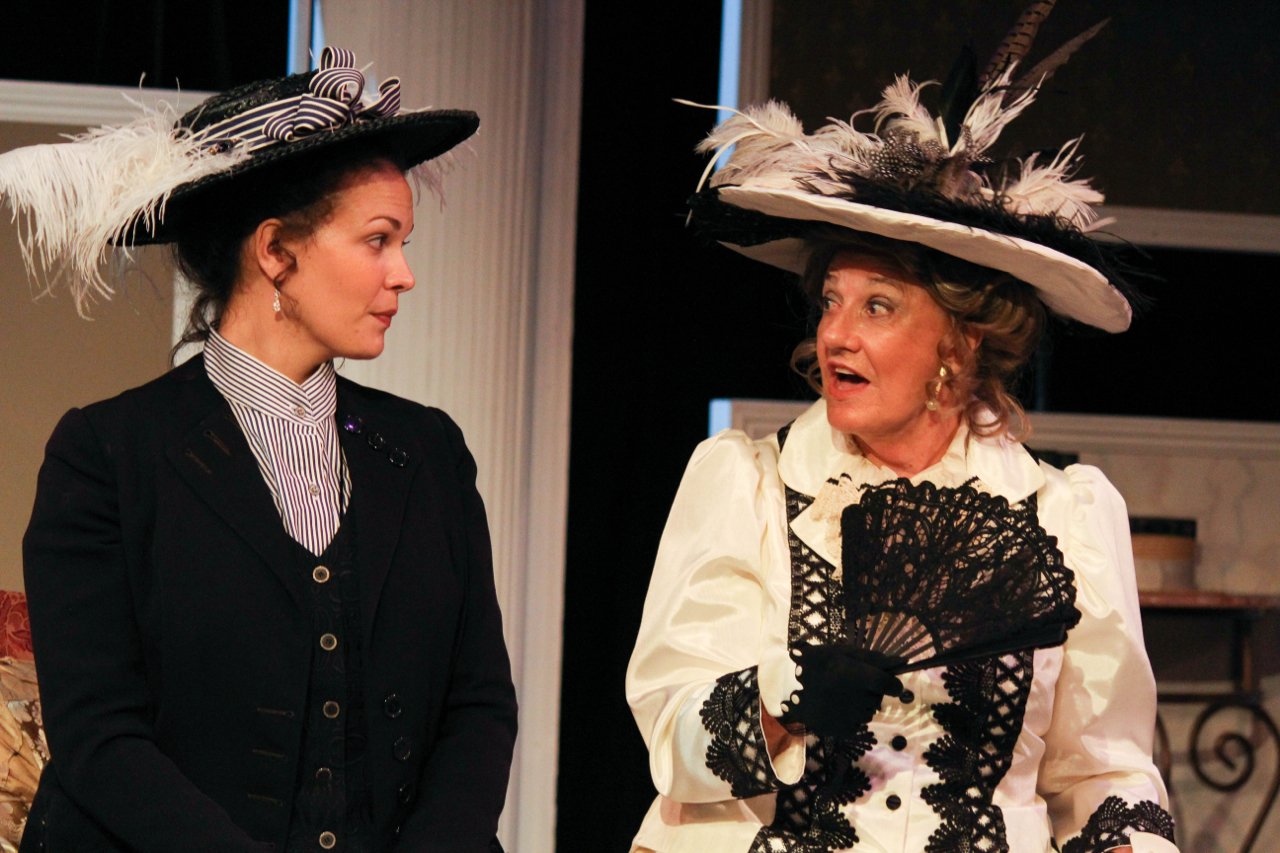 Two women in vintage costumes performing on stage, wearing elaborate hats with feathers, in a theatrical scene.