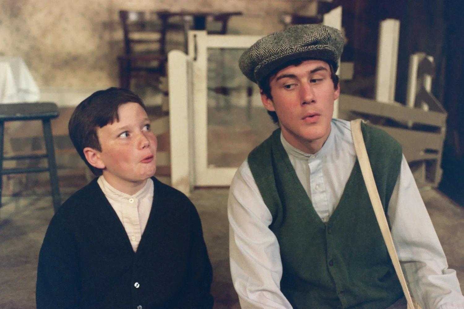 A young boy and an older boy seated indoors, with the older boy wearing a flat cap. The young boy is looking at the older boy with a surprised expression. The background shows a simple interior setting, possibly with chairs and a table.