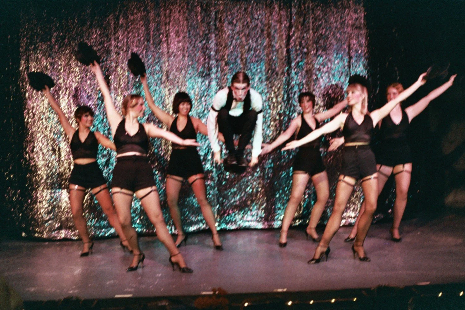 A group of dancers performing on stage in front of a sparkly curtain. The dancers are wearing black outfits, hats, and high heels, with one male dancer jumping in the center.