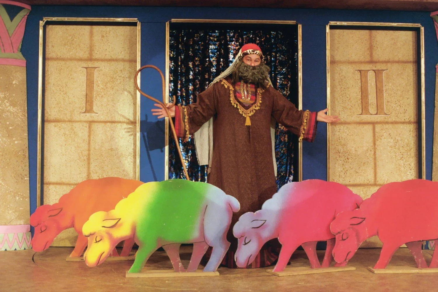 Stage performance with a person dressed in a biblical costume standing behind cutouts of colorful sheep.