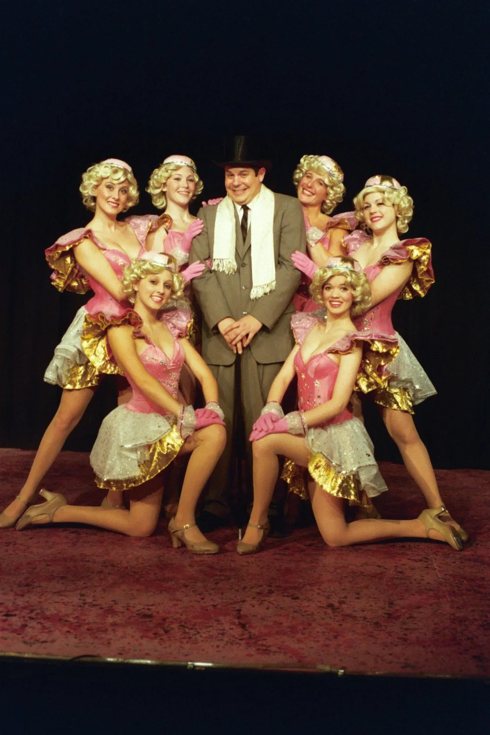 A man in a suit and top hat surrounded by six women in pink and gold stage costumes with blonde wigs, posing on stage.