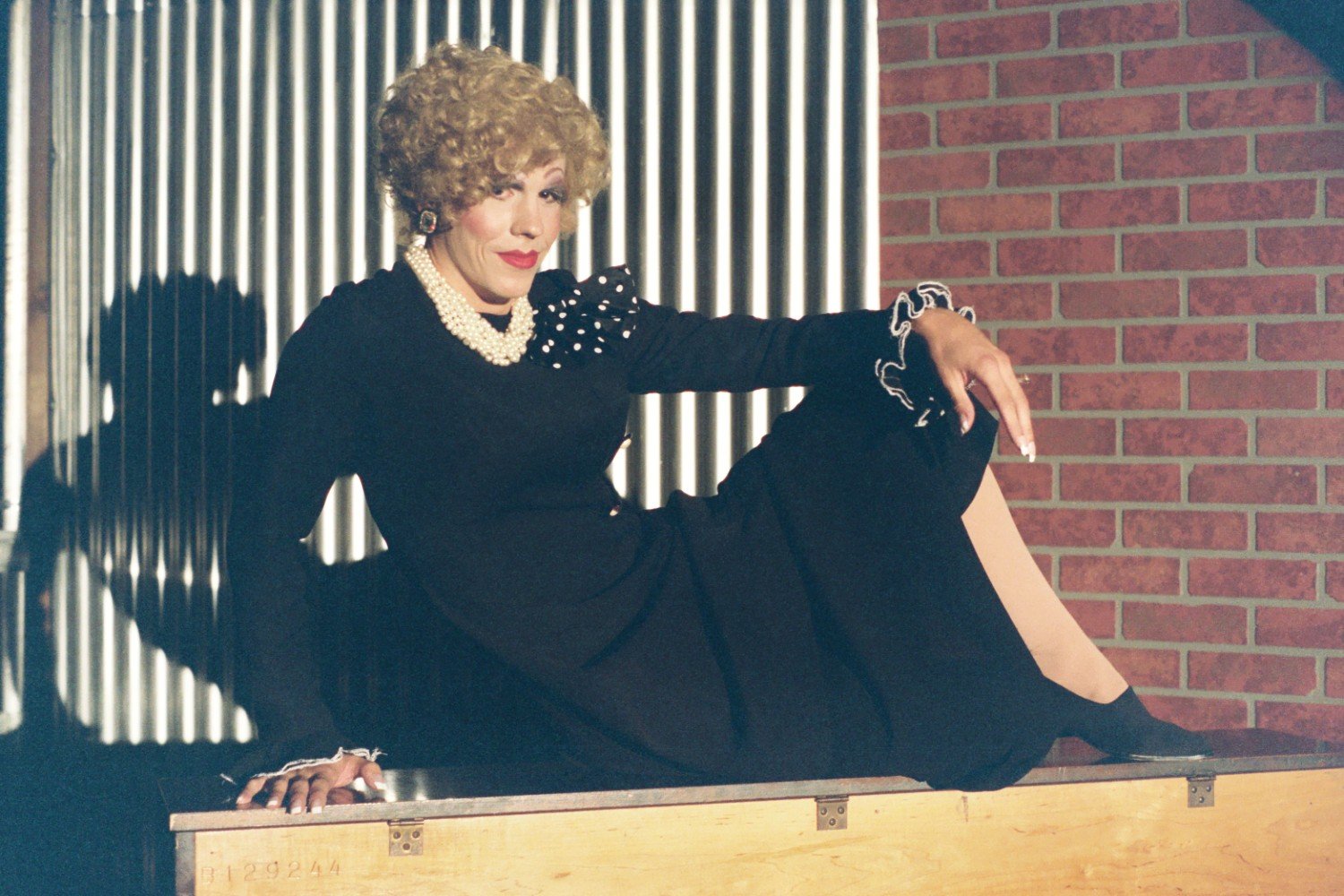 Person in black dress with pearl necklace sits on a wooden box in front of a brick and metal wall.