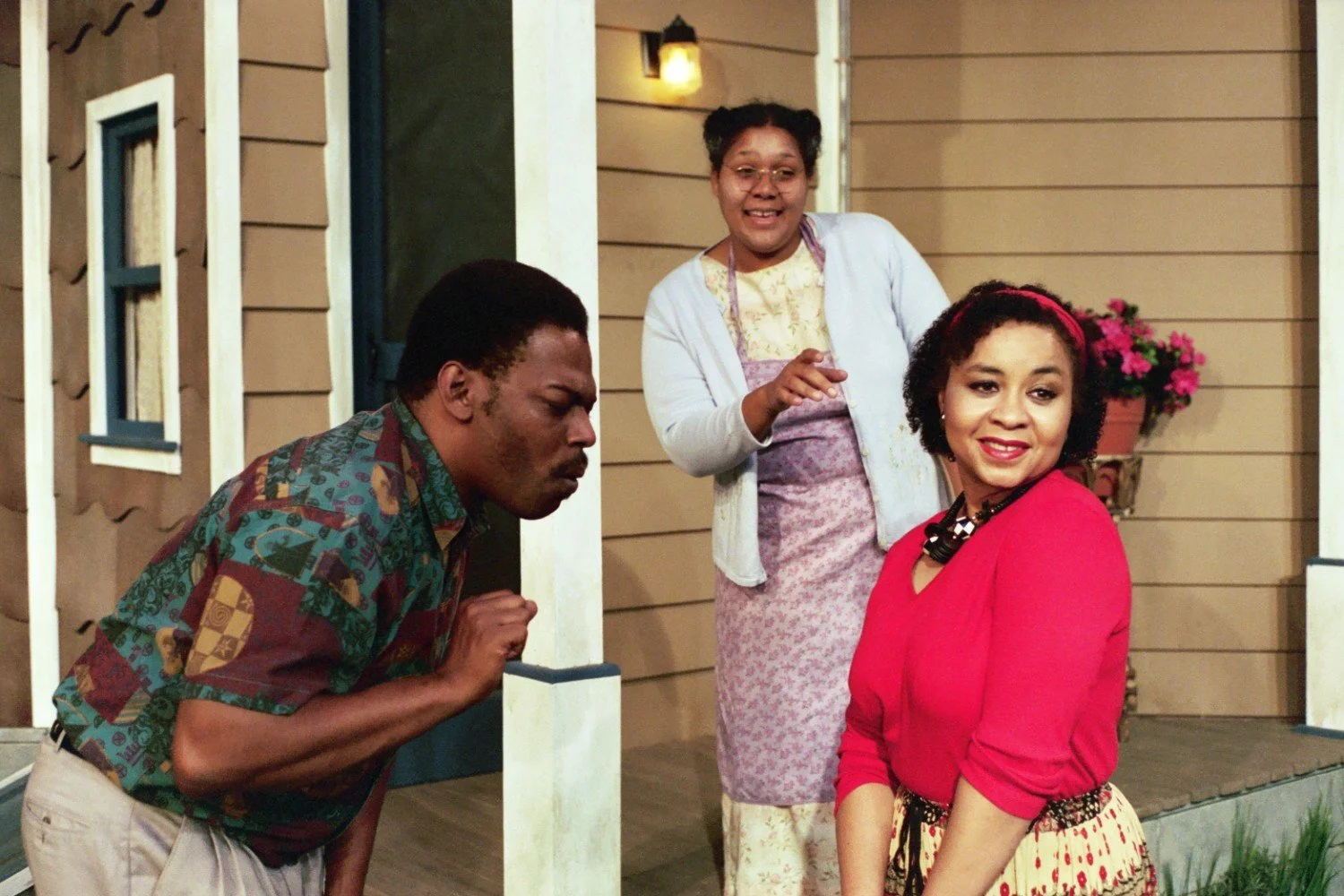 Three people on a porch, with one person in a patterned shirt, another in floral and lavender clothes, and one in a red top with a headband. The setting is outside a house with potted flowers.