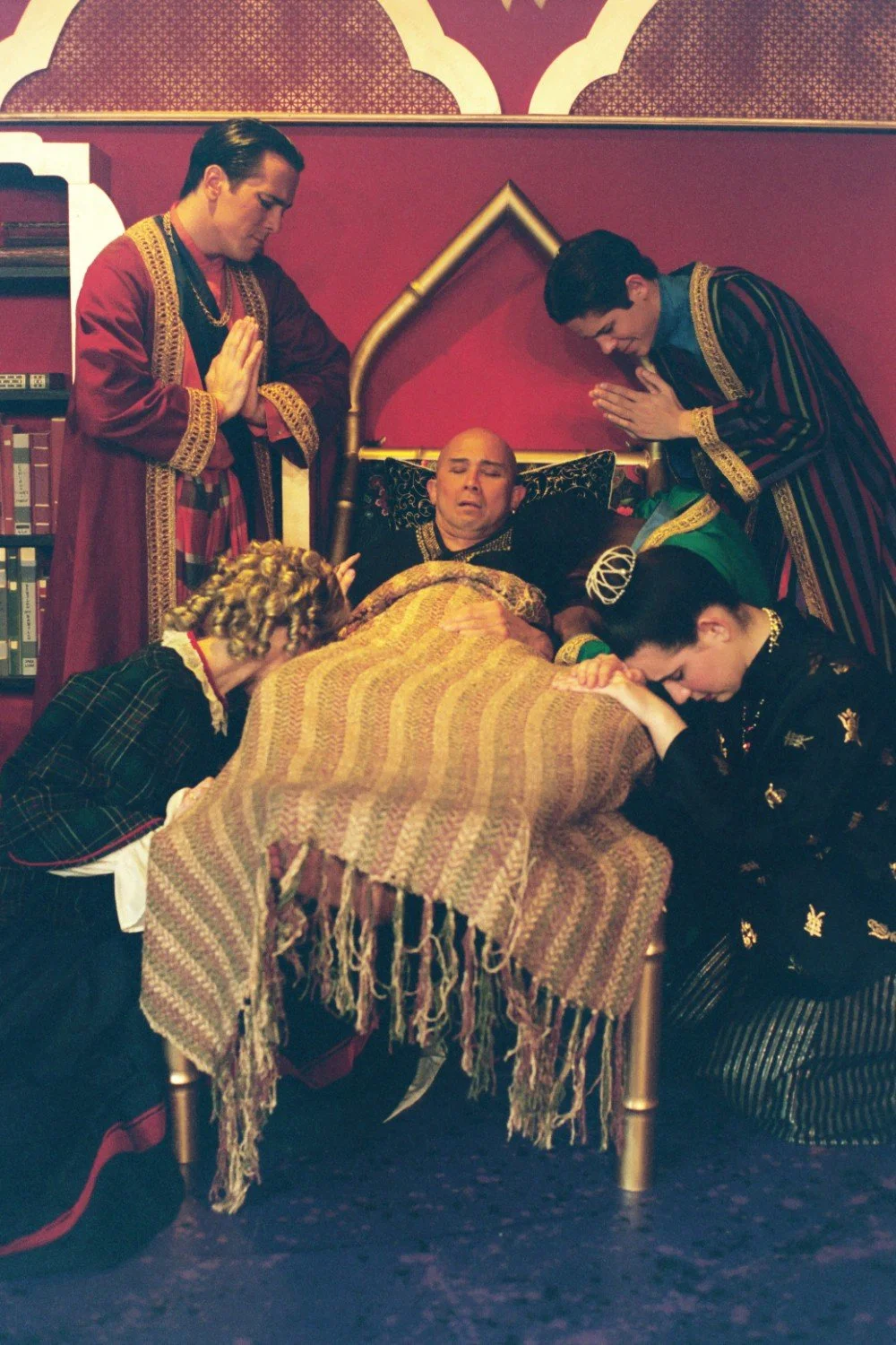 Dramatic scene with five people, one lying in bed covered with a blanket, while others stand or kneel in prayer or contemplation, set in a rich, theatrical setting with ornate costumes.