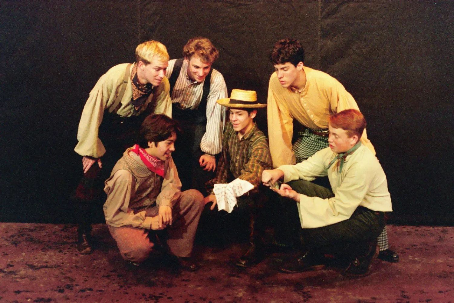 A group of six young men in period costumes, likely performing a scene from a play, gathered around a piece of paper one is holding. They appear engaged and collaborative. The setting suggests a theatrical production.