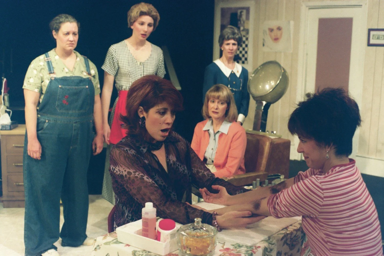 Scene from a theatrical performance with five women, one receiving a manicure.
