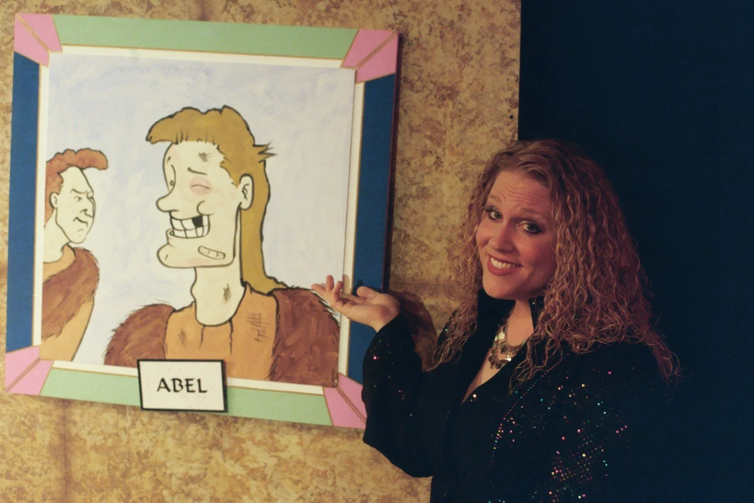 A woman with curly hair gestures towards a cartoon drawing of two cavemen named Abel, one smiling and the other frowning, on a decorated wall.