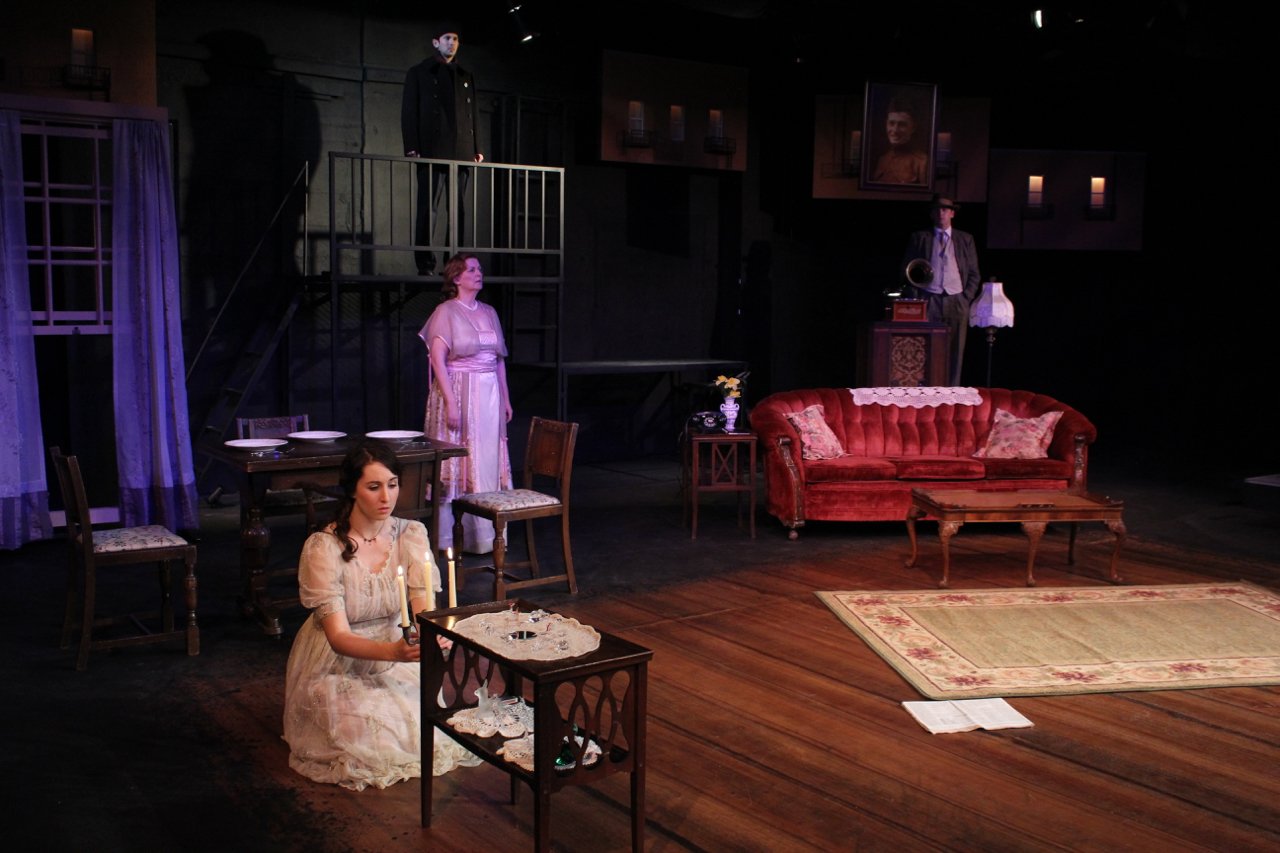 Theatrical stage set featuring a living room scene with a red sofa, wooden table, and chairs. A woman in a vintage dress kneels near a small table with candles. Other actors stand on a raised platform and in the background. Dim lighting enhances the