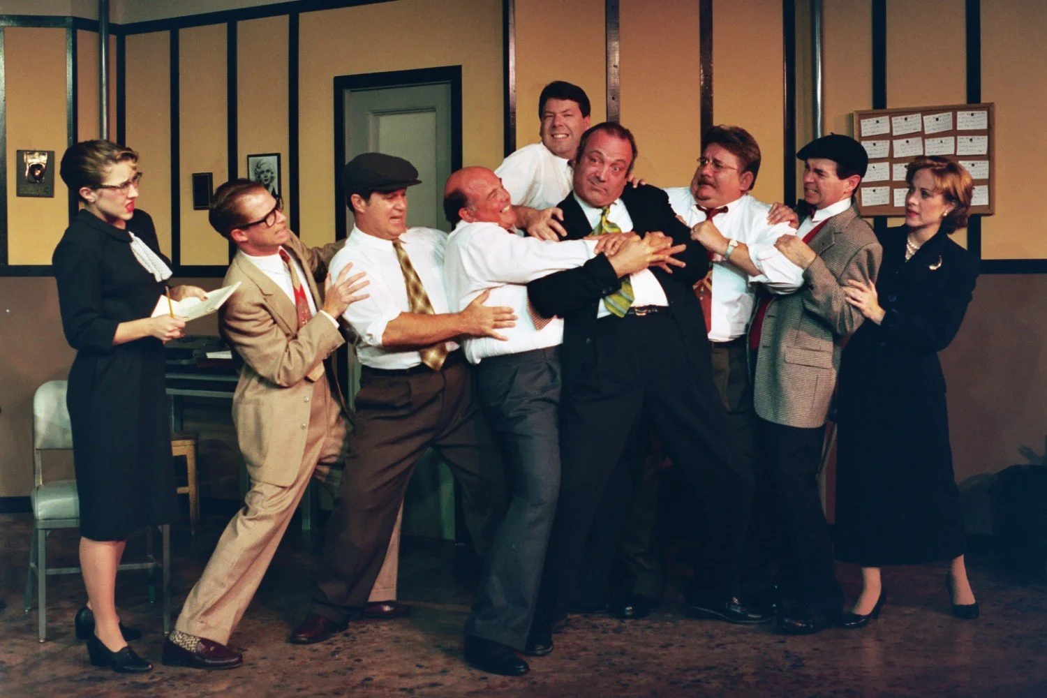 A group of people in period clothing engaged in a dramatic, theatrical scene inside a room. Eight men are physically interacting in a tense or comedic manner, while two women stand on the edges, one taking notes. The setting appears to be an office o