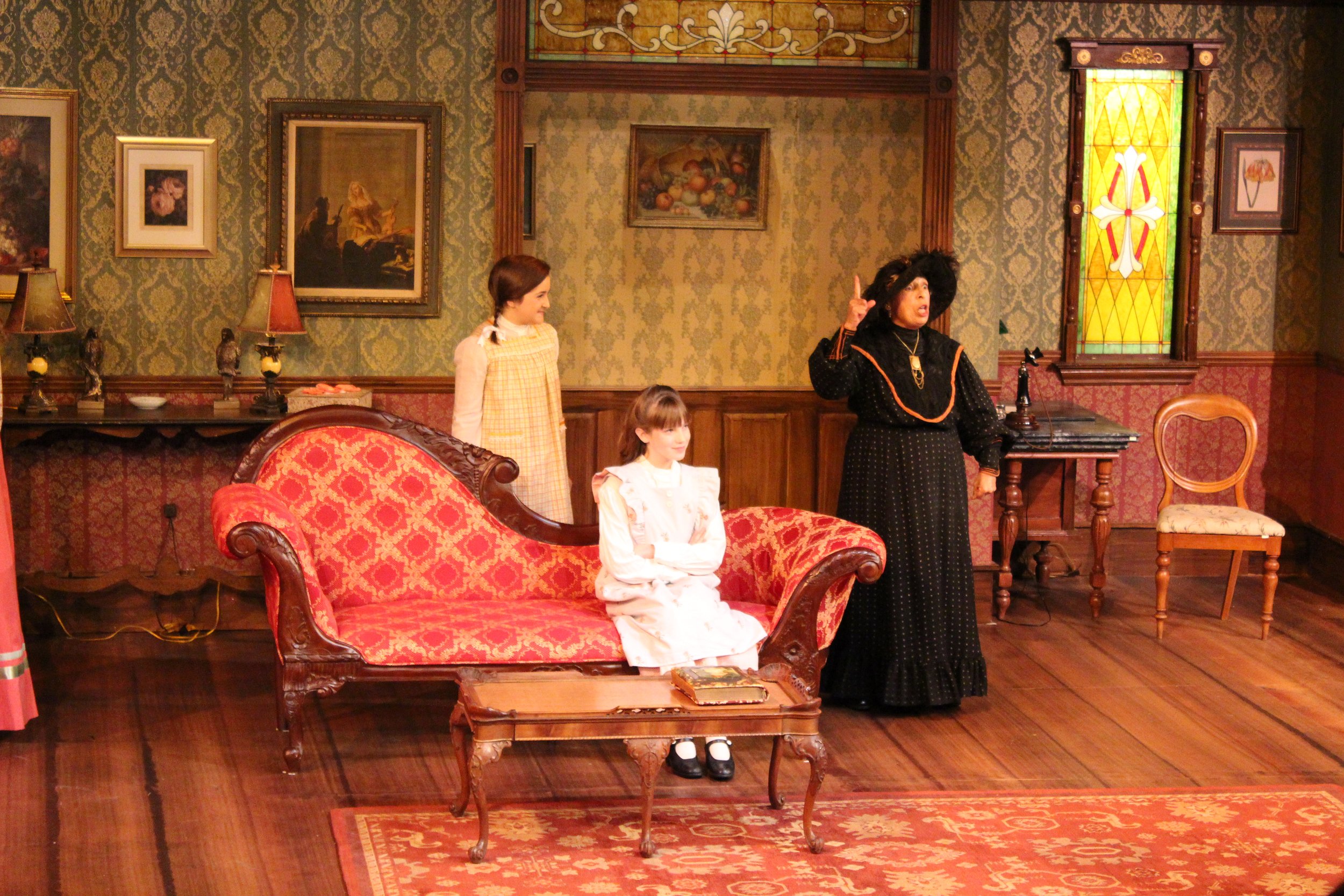 Three women in period costumes performing a stage play set in a vintage living room with ornate wallpaper, a red floral-patterned couch, and Victorian-style decor.