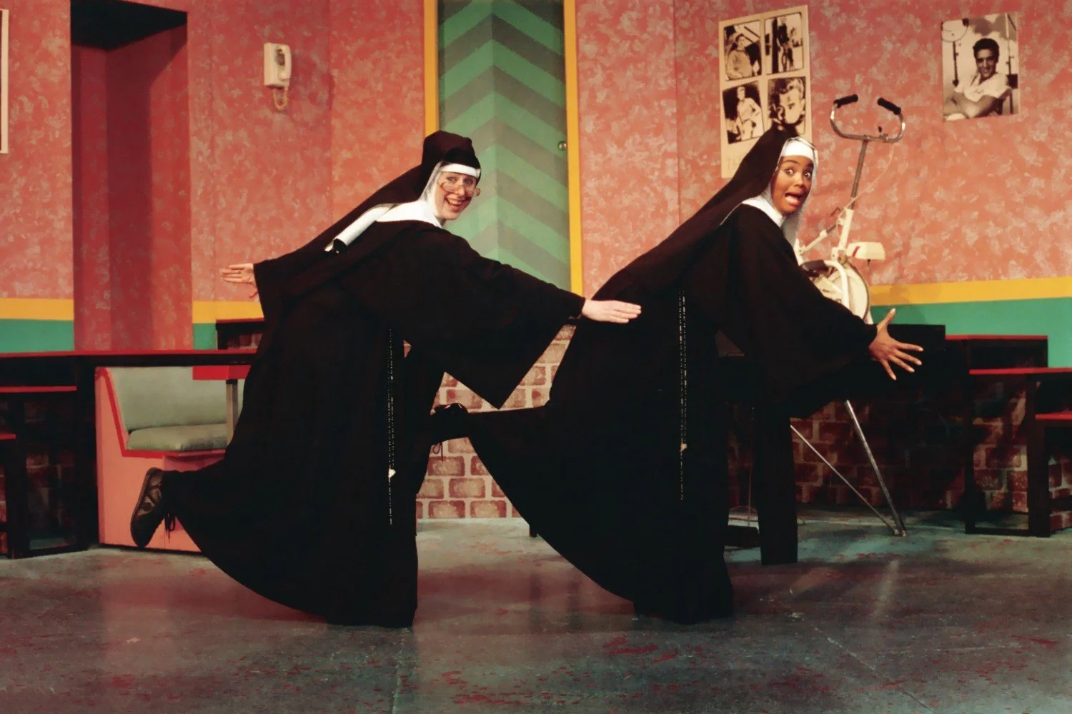 Two people in nun costumes performing a comedic dance in a colorful room, with exercise equipment and photos on the wall in the background.