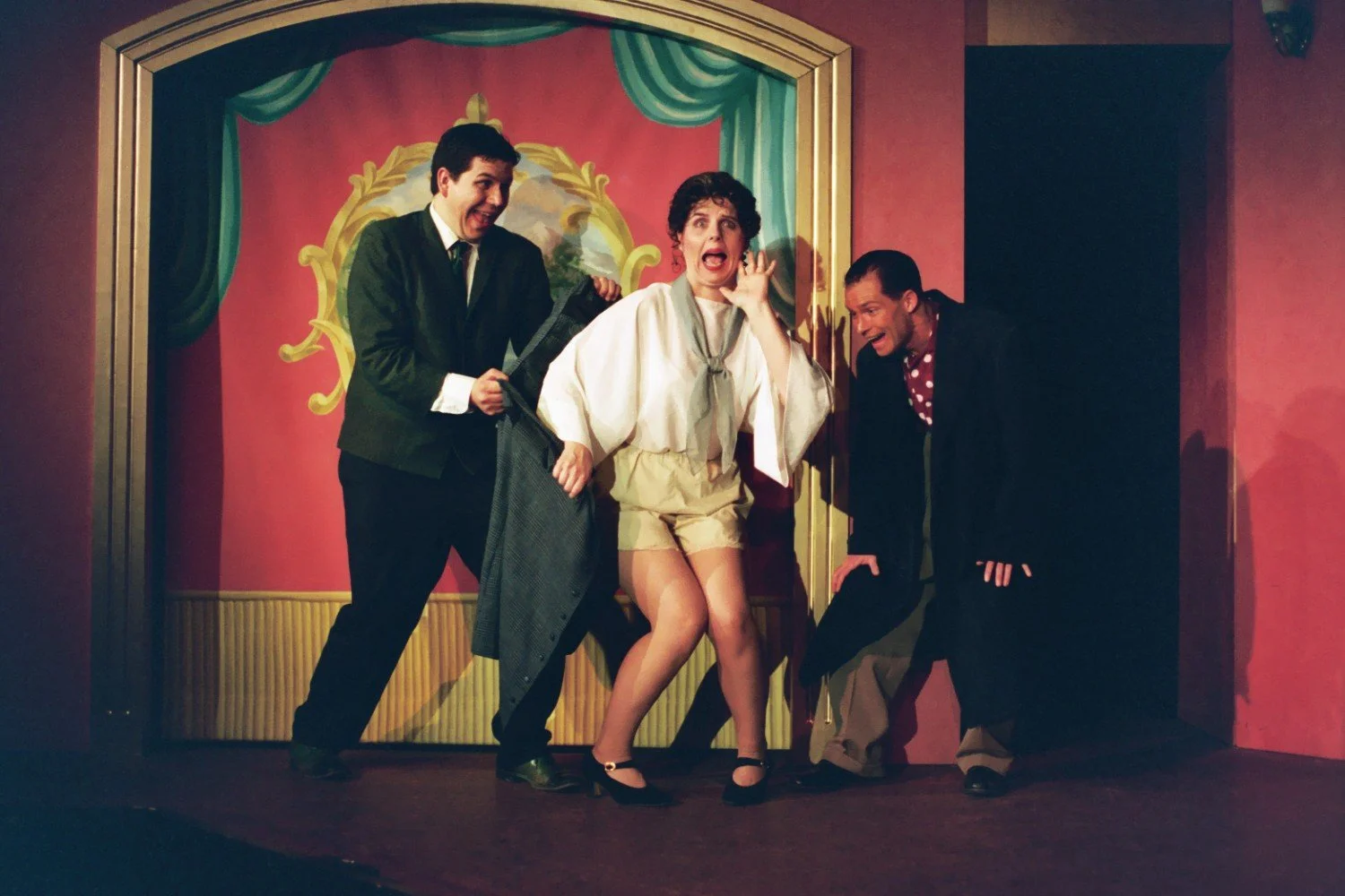 Three people performing a comedic scene on stage, one is in shorts, with surprised expressions against a colorful backdrop.