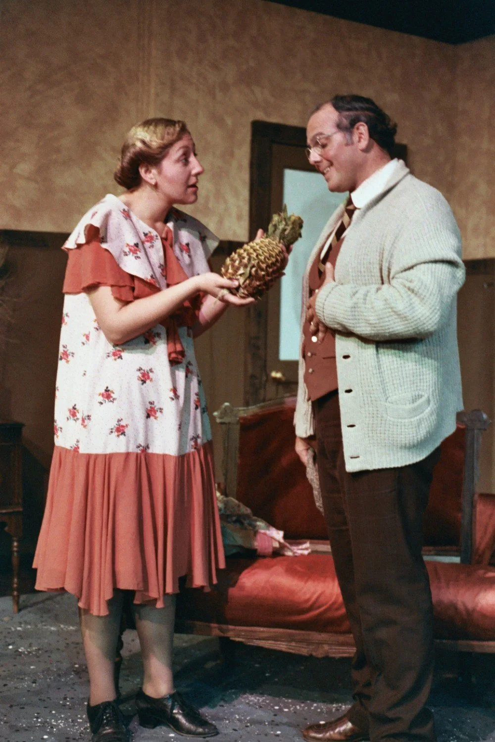 A woman holding a pineapple while talking to a man in a vintage setting.